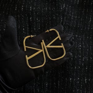 Puede incluir: Un par de pendientes de aro dorados con un dise&ntilde;o geom&eacute;trico. Los pendientes est&aacute;n hechos de metal y tienen un aspecto simple y moderno.