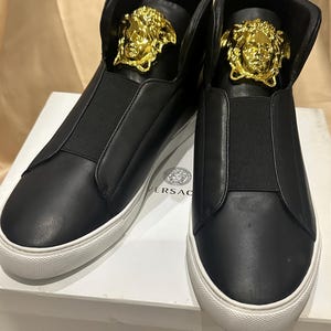 Op de afbeelding: Een paar zwarte high-top sneakers met witte zolen. Elke schoen heeft een goudkleurig Medusa-hoofd embleem op de tong. De sneakers hebben een slip-on ontwerp met een elastische band over de bovenkant.