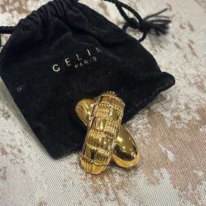 Pode incluir: Broche dourado com design entrançado e extremidades lisas e arredondadas. O broche está sobre uma superfície estampada junto a uma bolsa de veludo preta com "CELINE PARIS" impresso em dourado.