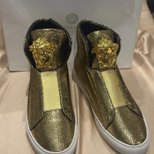 Op de afbeelding: Een paar gouden high-top sneakers met een textuurafwerking. De sneakers hebben een gouden Medusa-hoofdembleem en een witte zool. De schoenen worden gepresenteerd voor een witte doos met de merknaam.