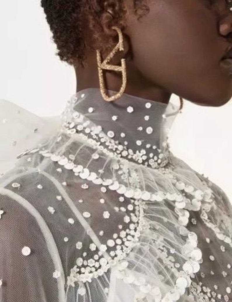 Puede incluir: Un top blanco de tul con cuello alto y cuello de volantes. El top est&aacute; adornado con lentejuelas y cuentas. La modelo lleva pendientes de aro dorados.