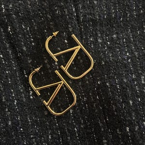 Puede incluir: Un par de pendientes de aro dorados con un dise&ntilde;o geom&eacute;trico. Los pendientes est&aacute;n hechos de metal grueso y tienen un aspecto simple y moderno.