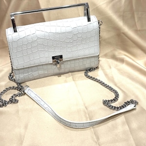 May include: White, textured crossbody bag with a silver chain strap and handle. The bag features a rectangular shape with a flap closure and a silver clasp. The bag's surface has a crocodile skin pattern.