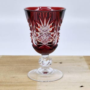 Könnte beinhalten: Ein rubinrotes Kristall-Weinglas mit detailliertem Sternmuster. Das Glas hat einen klaren, gedrehten Stiel und einen kreisförmigen Fuß. Das Glas steht auf einer hellen Holzoberfläche.