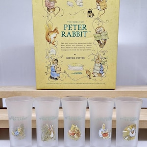 Puede incluir: Un juego de cinco vasos de vidrio esmerilado con ilustraciones de personajes de Peter Rabbit. Los vasos se exhiben frente a una caja vintage titulada "The World of Peter Rabbit" de Beatrix Potter.