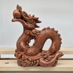 Könnte beinhalten: Eine detaillierte Drachenskulptur aus Terrakotta. Der Drache ist mit erhobenem Kopf dargestellt und hält eine Kugel im Maul. Die Skulptur weist komplizierte Schuppen, einen stacheligen Rückenkamm und dekorative Basiselemente auf.