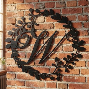 May include: Black metal wall art featuring a decorative wreath design with leaves, berries, and a heart accent. The letter "N" is centered within the wreath. The art is mounted on a red brick wall.