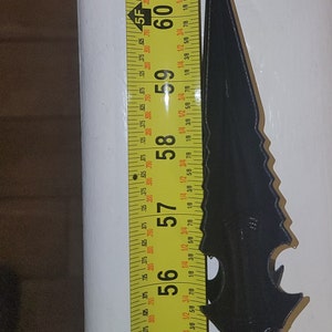 May include: A black metal knife with a serrated blade and a handle with a textured design. The knife is measured against a yellow tape measure that reads 54 to 60 inches.