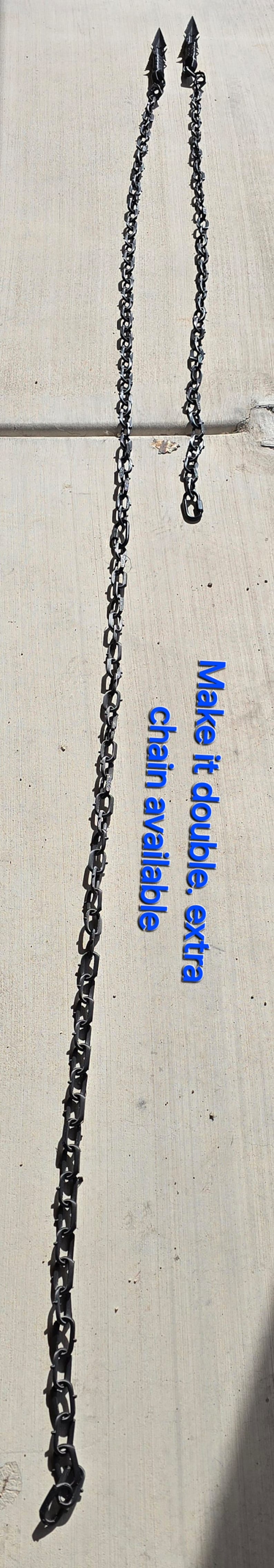 May include: Two black metal chains with sharp points at the ends. The chains are hanging straight down on a light gray concrete surface. The text "Make it a double. Extra chain available." is written in blue letters on the concrete.