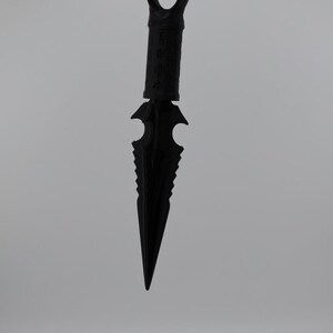May include: A black metal dagger with a serrated blade and a ring at the top. The dagger is hanging from a chain.