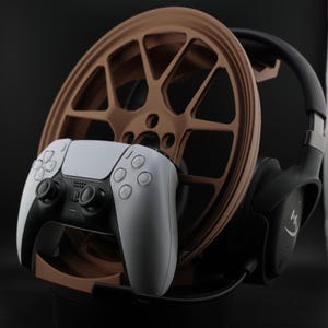 May include: A black and white video game controller and black headphones are displayed on a brown and black stand. The stand is shaped like a car wheel rim.