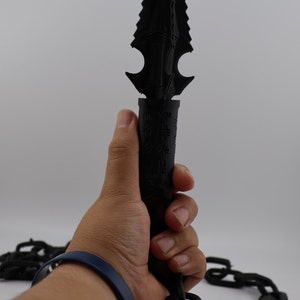 May include: A black, detailed, spiky dagger with a chain attached to the handle. The dagger is being held by a hand wearing a blue bracelet.