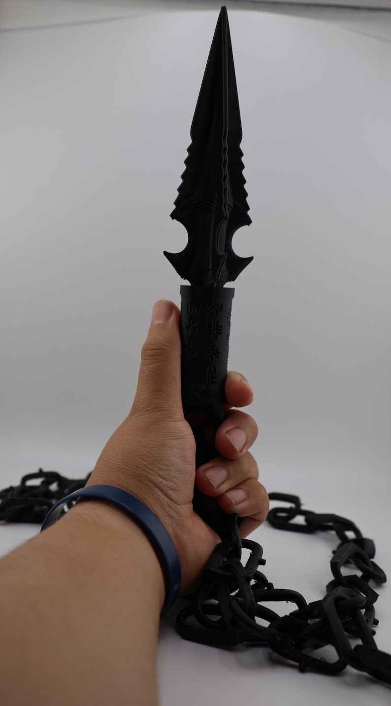 May include: A black, detailed, spiky dagger with a chain attached to the handle. The dagger is being held by a hand wearing a blue bracelet.