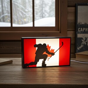 May include: A rectangular light box displaying the Canadian flag, with a silhouette of a hockey player. The flag's red and white design is illuminated, and the hockey player is in black. The light box is on a wooden surface.