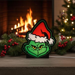 May include: A green Grinch Christmas decoration, featuring a red Santa hat with white trim. The Grinch's face has yellow eyes and a black outline. The decoration is on a stone surface, with a fireplace and Christmas tree in the background.