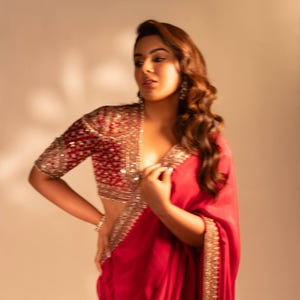 Op de afbeelding: Een vrouw draagt een rode sari met gouden details en een bijpassende rode blouse met gouden versieringen. De blouse heeft korte mouwen en een diepe V-hals. De sari is elegant gedrapeerd, waardoor zachte plooien ontstaan.