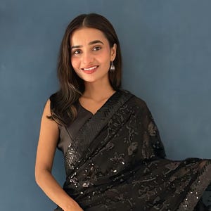 May include: A black saree with intricate sequin and floral embroidery. The sleeveless top complements the saree's design. The saree is draped elegantly, showcasing its texture and sheen. The background is a solid blue wall.