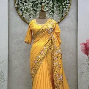 May include: A yellow saree with intricate embroidery featuring floral and bird motifs. The saree is draped on a mannequin against a white background with a floral backdrop.