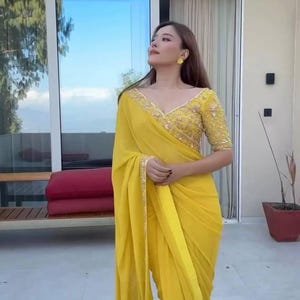 Pu&ograve; includere: Una donna indossa un sari giallo brillante con ricami dorati intricati sul corpetto e sul bordo. Il sari &egrave; drappeggiato con eleganza, creando morbide pieghe. L'outfit include orecchini gialli abbinati.