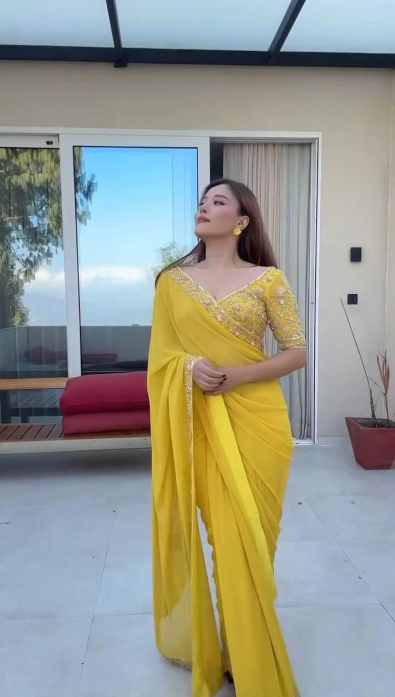 Pu&ograve; includere: Una donna indossa un sari giallo brillante con ricami dorati intricati sul corpetto e sul bordo. Il sari &egrave; drappeggiato con eleganza, creando morbide pieghe. L'outfit include orecchini gialli abbinati.
