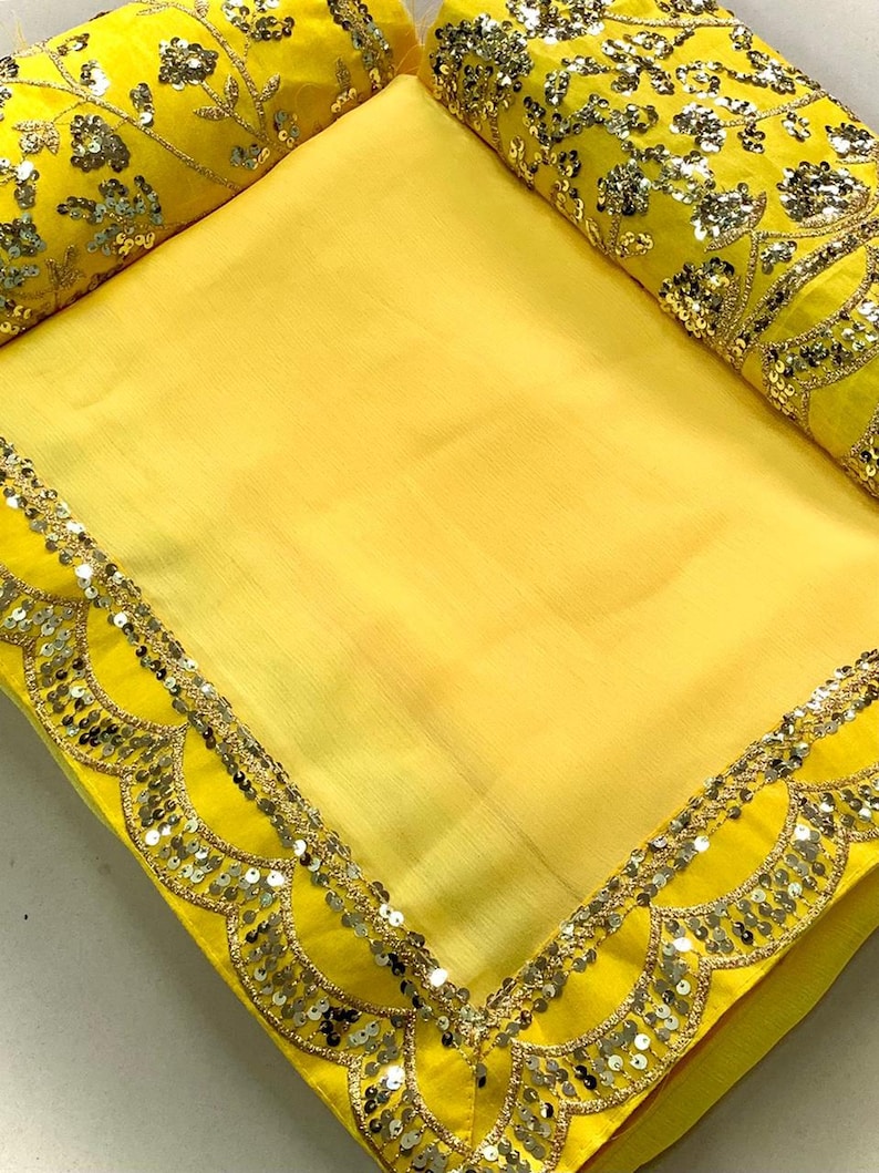 Pu&ograve; includere: Un sari giallo brillante con un bordo smerlato impreziosito da paillettes argentate e finiture dorate. Il sari ha un pezzo di camicetta giallo abbinato con intricati ricami floreali e dettagli di paillettes. Il tessuto sembra essere un materiale leggero e fluido.