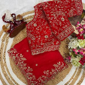 Puede incluir: Un vibrante conjunto de sari rojo con intrincados bordados dorados. El conjunto incluye una blusa y una tela drapeada, ambas adornadas con detallados diseños florales y de cachemira. El conjunto se exhibe sobre una estera de fibra natural, mostrando las ricas texturas y colores.