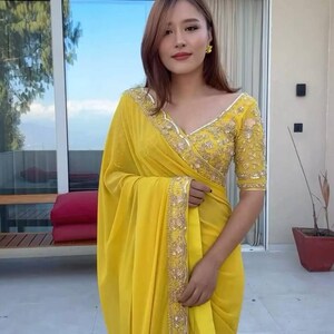 Pu&ograve; includere: Una donna indossa un sari giallo brillante con ricami dorati. Il sari ha una camicetta con scollo a V con maniche corte e dettagli dorati intricati. Lo sfondo include una finestra e una pianta in vaso.