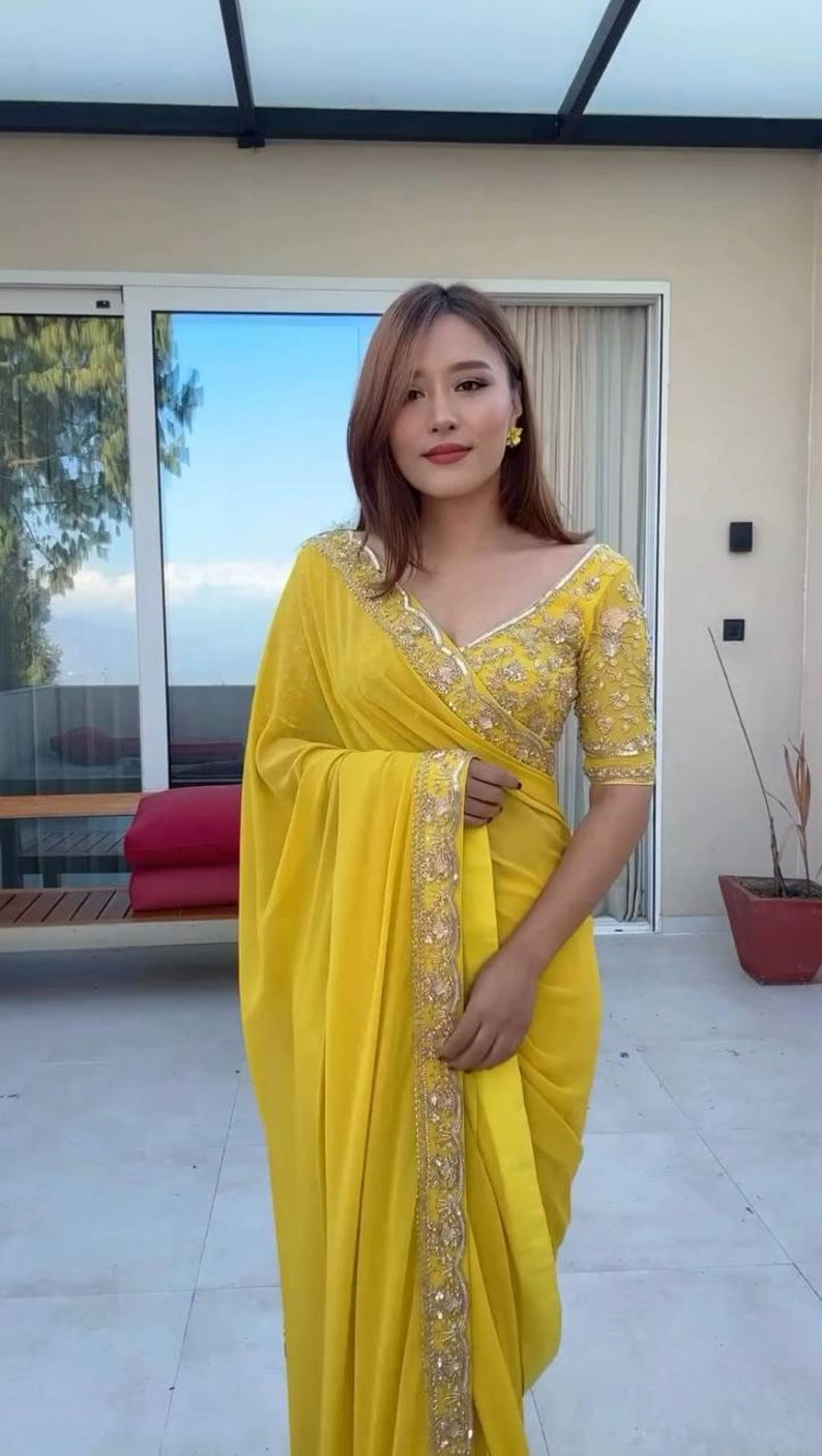 Pu&ograve; includere: Una donna indossa un sari giallo brillante con ricami dorati. Il sari ha una camicetta con scollo a V con maniche corte e dettagli dorati intricati. Lo sfondo include una finestra e una pianta in vaso.