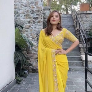 Pu&ograve; includere: Una donna indossa un sari giallo brillante con ricami dorati intricati. Il sari &egrave; drappeggiato con eleganza, con una blusa abbinata con dettagliate perline. Lo sfondo include muri e scale in pietra.