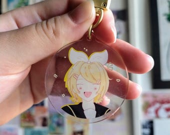 May include: A clear, round keychain with a gold-colored clasp. The keychain features a cartoon illustration of a smiling character with blonde hair, a white bow, and a black jacket. The background is a collection of colorful images.