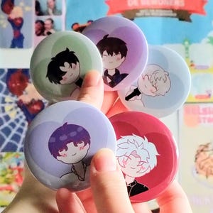 May include: Five circular badges are held in a hand, each displaying an anime-style character. The characters have various hair colors, such as black, purple, and white, and are set against pastel backgrounds of green, purple, blue, and red.