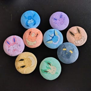 May include: A collection of ten round, colorful buttons featuring various cartoon character designs. The buttons are in shades of blue, purple, pink, orange, and green, with detailed illustrations of the characters. The buttons are arranged on a dark surface.