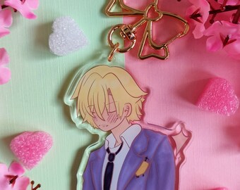 May include: A clear acrylic charm featuring an anime-style character with blonde hair, wearing a purple blazer and black tie. The charm has a gold bow-shaped clasp. The background is split between pink and light green, with pink flower and heart decorations.