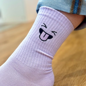 Könnte beinhalten: Ein Paar lavendelfarbene Socken mit einem schwarz-weißen Smiley, der die Zunge herausstreckt.
