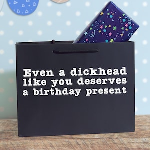 May include: A black gift bag with the white text "Even a dickhead like you deserves a birthday present" printed on the front. A rectangular gift box wrapped in dark blue paper with a colourful star and confetti pattern is inside the bag.