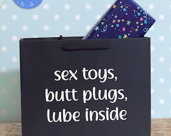 May include: A black gift bag with the white text "sex toys, butt plugs, lube inside" printed on the front. A rectangular gift box wrapped in dark blue paper with a colourful pattern of stars and confetti is tucked inside the bag.