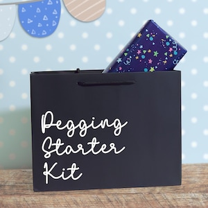 May include: A black gift bag with the white text Pegging Starter Kit printed on the front. A rectangular box wrapped in dark blue paper with a colourful pattern of small stars, circles, and confetti shapes is tucked inside the bag.