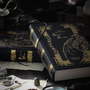 Op de afbeelding: Twee zwarte hardcover boeken met gouden accenten en ingewikkelde ontwerpen. Het bovenste boek heeft de titel "The Empyrean Iron Flame" en de auteur "Rebecca Yarros". Het onderste boek heeft de titel "The Fourth Wing".