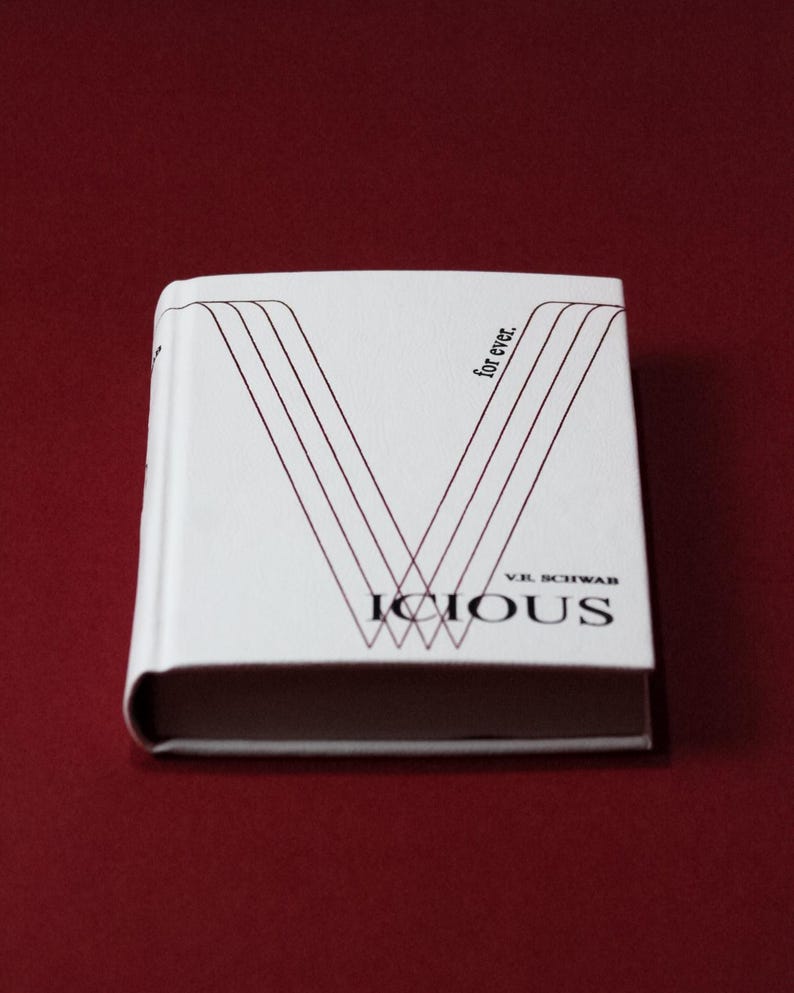 May include: A hardcover book titled "Vicious" by V.E. Schwab. The book has a white cover with a stylized "V" design in burgundy lines. The words "for ever." are printed vertically on the cover.