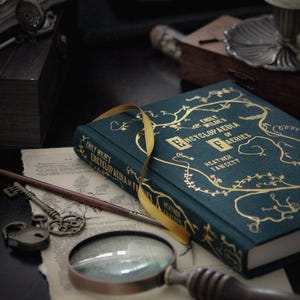 May include: A dark green hardcover book with gold lettering titled "Emily Wilde's Encyclopedia of Faeries" by Heather Fawcett. The book is lying open on a table with a magnifying glass, a key, and a piece of paper with writing on it.