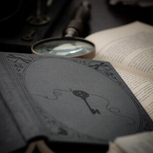 May include: An open book with a dark gray cover featuring a key illustration. A magnifying glass and other books are in the background, creating a vintage, antique aesthetic. The image has a moody, mysterious feel.