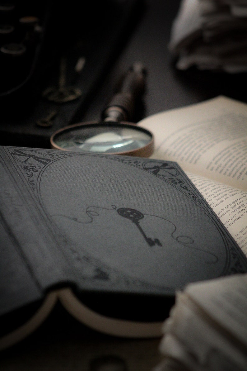 May include: An open book with a dark gray cover featuring a key illustration. A magnifying glass and other books are in the background, creating a vintage, antique aesthetic. The image has a moody, mysterious feel.
