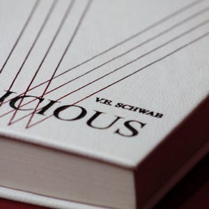 May include: A hardcover book with a white cover and the title "Vicious" in large black letters. The author's name, V.E. Schwab, is printed below the title. Red lines extend from the top of the cover. The book is set against a dark red background.