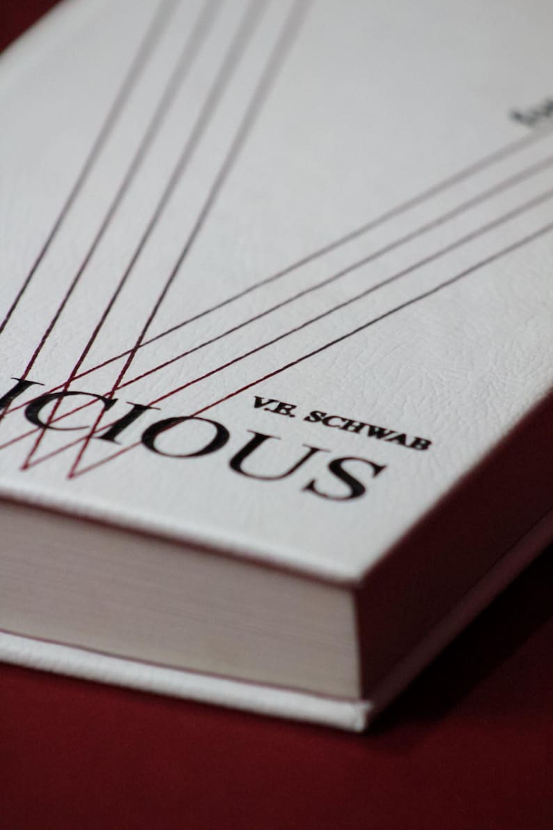 May include: A hardcover book with a white cover and the title "Vicious" in large black letters. The author's name, V.E. Schwab, is printed below the title. Red lines extend from the top of the cover. The book is set against a dark red background.