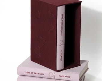 Puede incluir: Un estuche para libros de color granate oscuro con patrones de corazones en relieve, que contiene un libro rosa claro titulado Love, Theoretically de Hazelwood. Dos libros rosa claro adicionales, Love on the Brain y The Love Hypothesis de Hazelwood, están apilados debajo.
