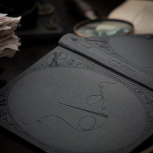 May include: A close-up of a dark gray notebook with a needle and thread design. The notebook has an ornate border and the word "coraline" is visible. Other items in the background include a magnifying glass and a stack of papers.