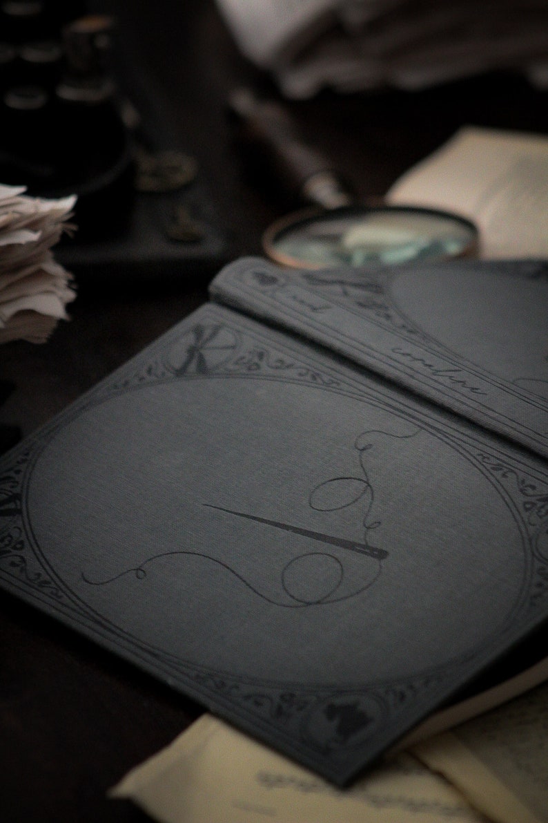 May include: A close-up of a dark gray notebook with a needle and thread design. The notebook has an ornate border and the word "coraline" is visible. Other items in the background include a magnifying glass and a stack of papers.