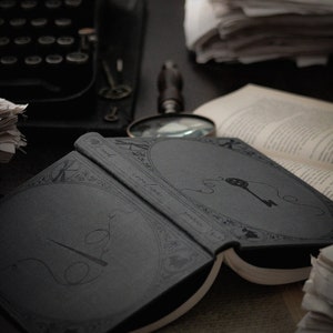 May include: An open, dark gray book with decorative line art of a key and needle and thread. A magnifying glass, typewriter, and stacks of paper are in the background. The book's pages are off-white.