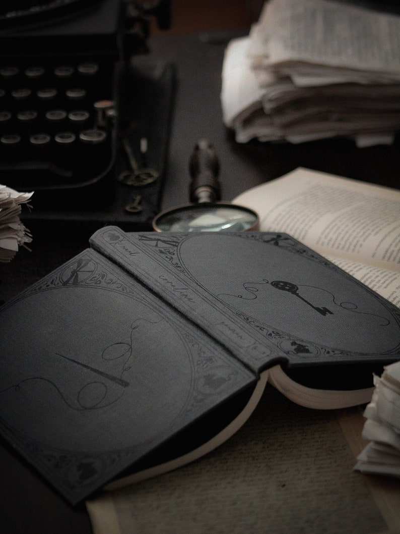May include: An open, dark gray book with decorative line art of a key and needle and thread. A magnifying glass, typewriter, and stacks of paper are in the background. The book's pages are off-white.