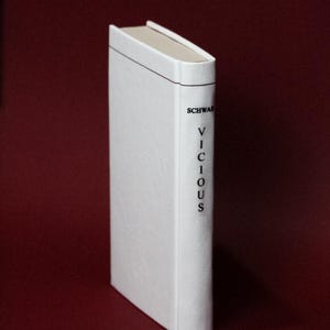 May include: A hardcover book with a white cover and the title "VICIOUS" printed vertically in black. The author's name, "SCHWAB", is printed above the title. The book is set against a dark red background.